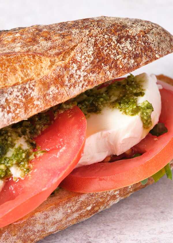 Close-up view of a caprese sandwich with fresh slices of tomato, thick cuts of mozzarella cheese, and vibrant green pesto, served on a crusty bread roll dusted with flour.