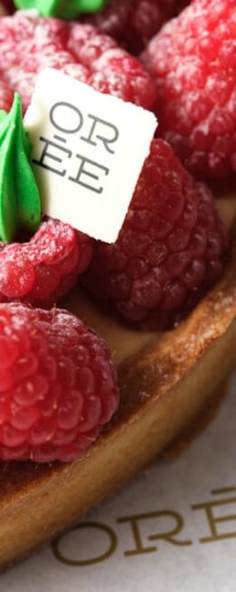 Gourmet raspberry tart filled with creamy vanilla custard, topped with fresh, dusted raspberries and a decorative green icing swirl on a white chocolate square bearing the letters 'OREE'.