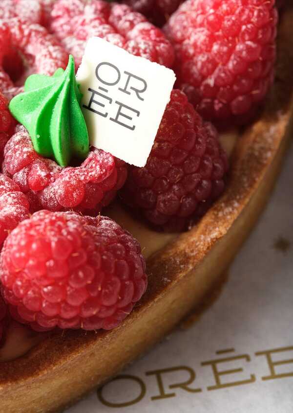 Gourmet raspberry tart filled with creamy vanilla custard, topped with fresh, dusted raspberries and a decorative green icing swirl on a white chocolate square bearing the letters 'OREE'.