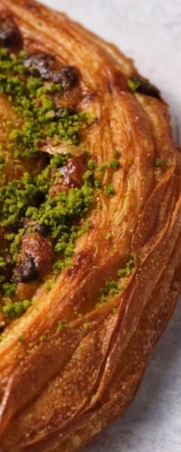 Close-up of a flaky, golden pastry topped with vibrant green pistachio crumbs, showcasing its layered texture and sugary glaze.