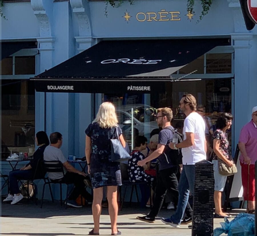 A bustling outdoor scene at ORÉE Boulangerie and Patisserie, showing customers enjoying their meals and others waiting to be served. The café, with its elegant black and blue façade, invites a mix of people under its shaded awning, reflecting a vibrant urban morning.