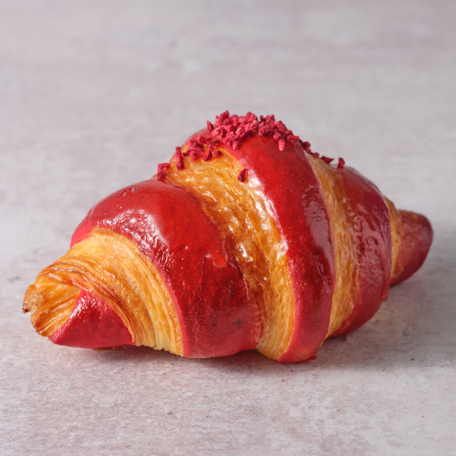 An exquisite croissant with a vibrant red glaze, featuring a perfectly layered and flaky texture that shines through the glossy exterior. The croissant curves elegantly, highlighting the meticulous craftsmanship and unique coloration.