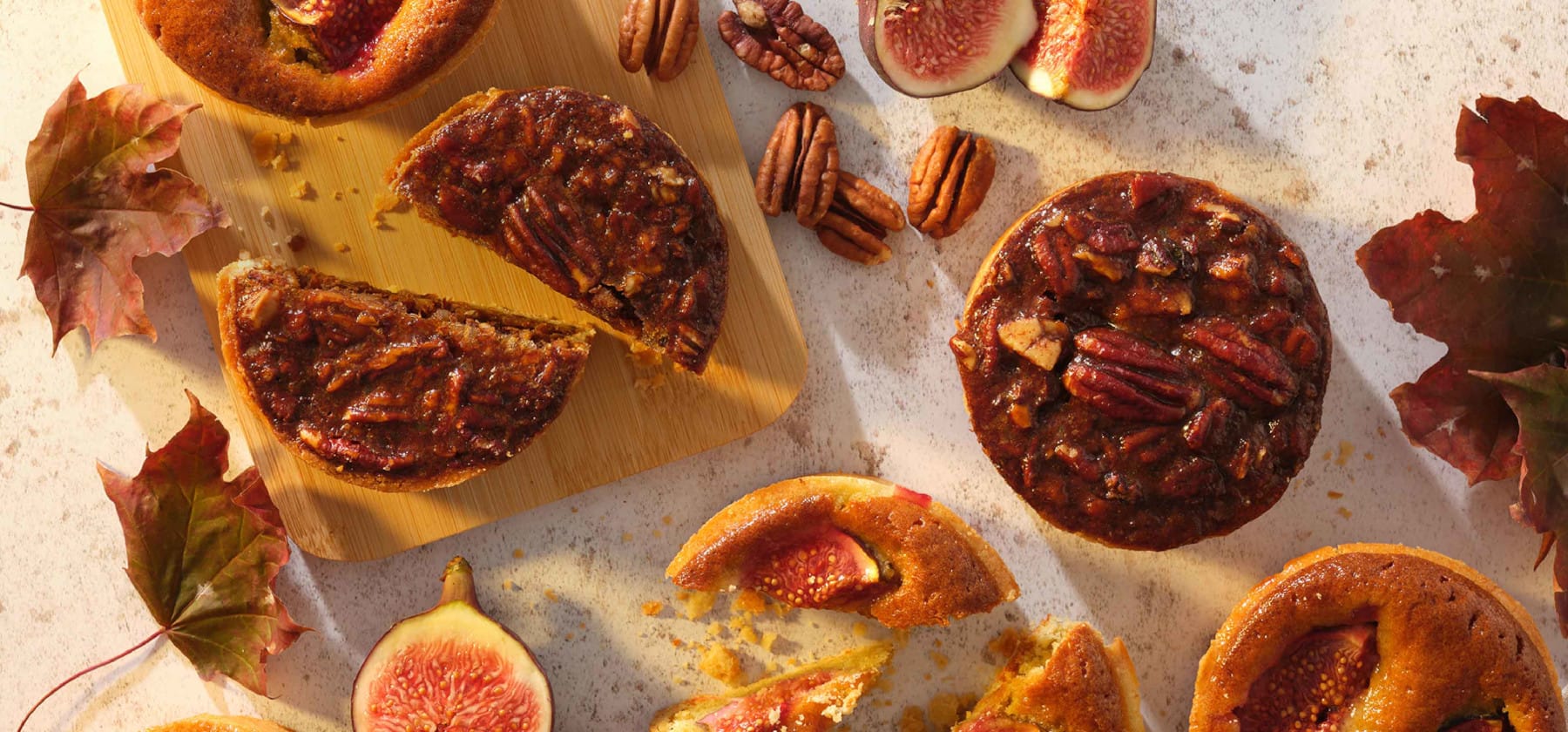 Assortment of autumn-inspired desserts featuring pecan tarts and fig pastries on a wooden cutting board, surrounded by scattered pecans, fresh fig halves, and colourful autumn leaves. The warm lighting enhances the golden and caramel tones of the sticky, candied pecans and the vibrant reds of the fig interiors, creating an inviting seasonal display.