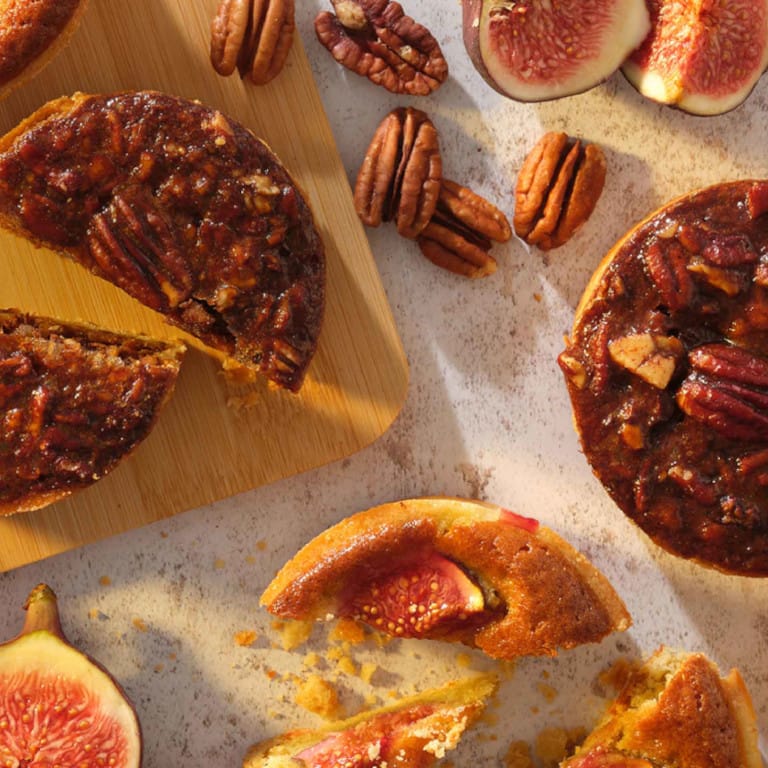 Assortment of autumn-inspired desserts featuring pecan tarts and fig pastries on a wooden cutting board, surrounded by scattered pecans, fresh fig halves, and colourful autumn leaves. The warm lighting enhances the golden and caramel tones of the sticky, candied pecans and the vibrant reds of the fig interiors, creating an inviting seasonal display.