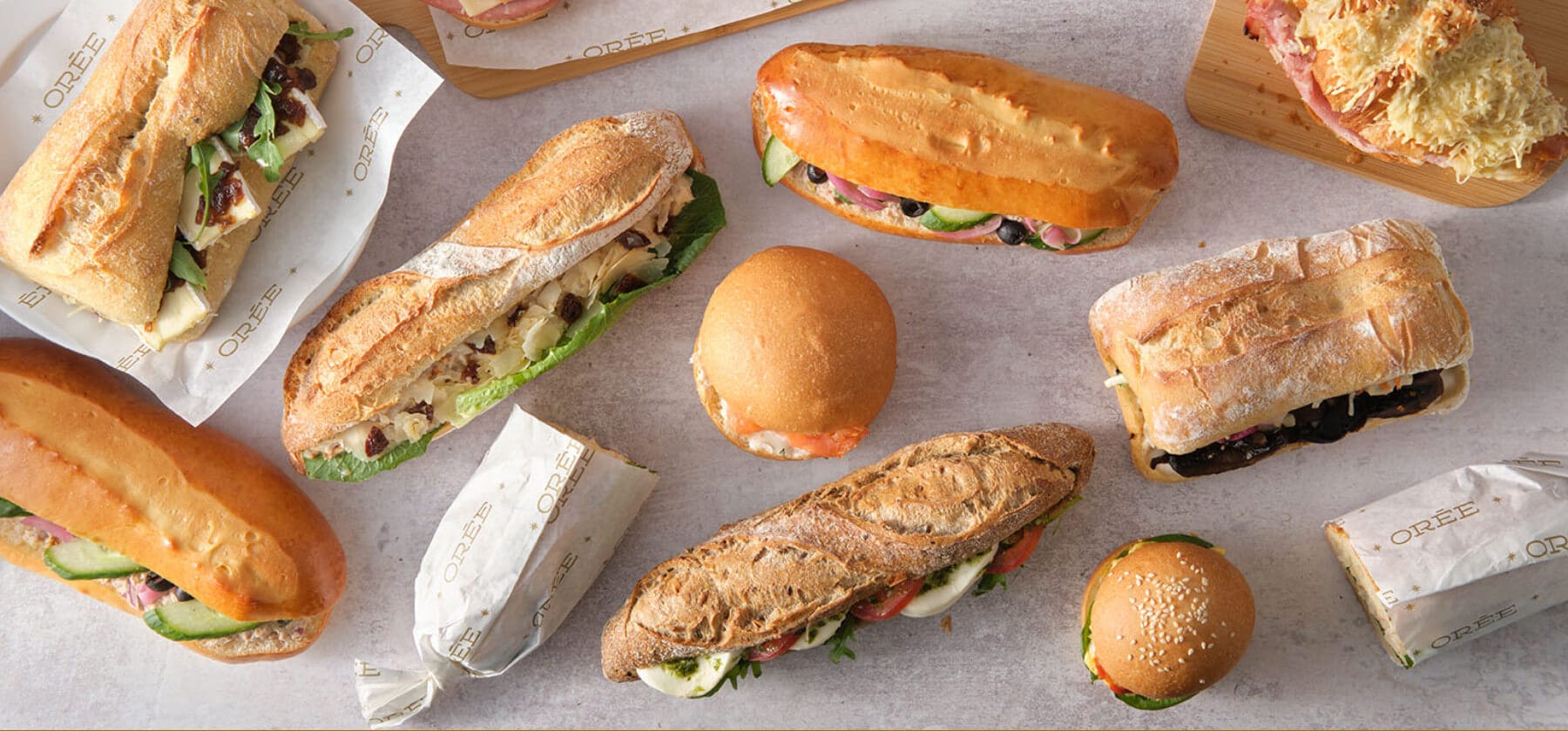 Assortment of gourmet sandwiches from OREE, featuring a variety of artisan breads including baguettes, rolls, and loaves. Each sandwich is filled with fresh ingredients like leafy greens, cheeses, and meats, and is presented on branded parchment paper. The display showcases the sandwiches in a neat and appetising arrangement.