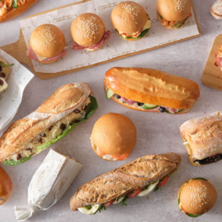 Assortment of gourmet sandwiches from OREE, featuring a variety of artisan breads including baguettes, rolls, and loaves. Each sandwich is filled with fresh ingredients like leafy greens, cheeses, and meats, and is presented on branded parchment paper. The display showcases the sandwiches in a neat and appetising arrangement.