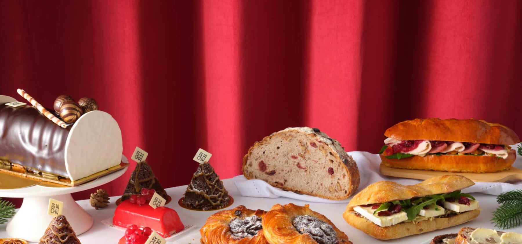 Festive Christmas bakery display featuring a chocolate Yule log, seasonal pastries, artisan bread, and sandwiches filled with festive ingredients, set against a rich red curtain backdrop.