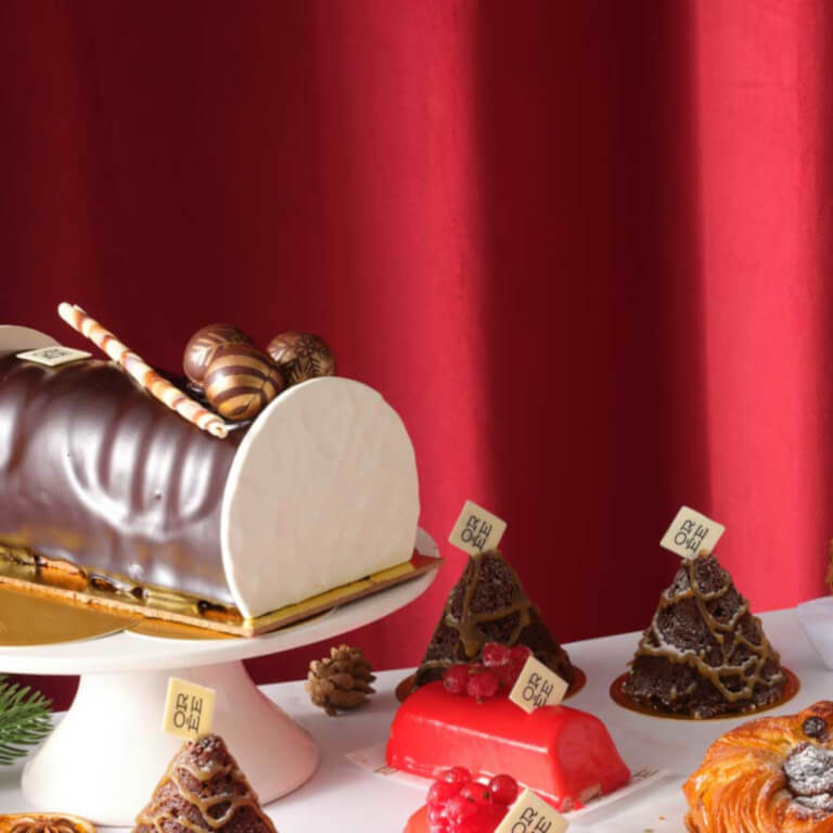 Festive Christmas bakery display featuring a chocolate Yule log, seasonal pastries, artisan bread, and sandwiches filled with festive ingredients, set against a rich red curtain backdrop.