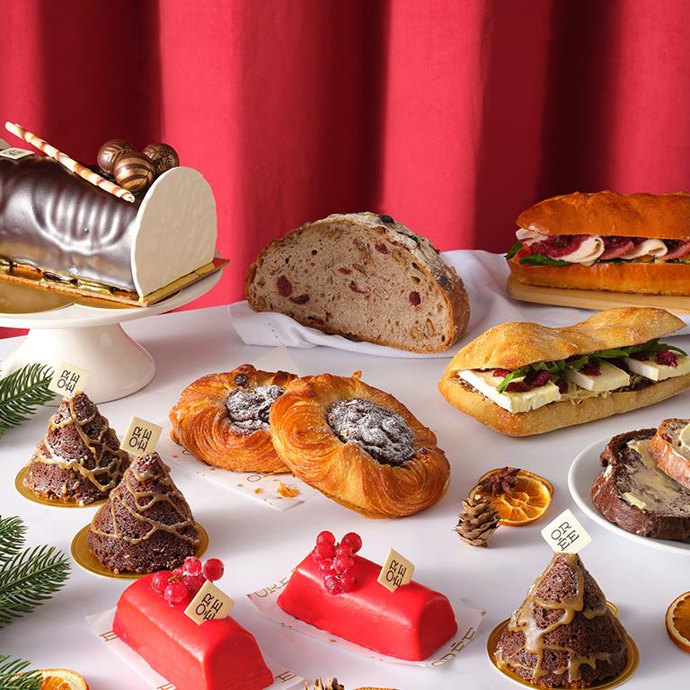 Festive Christmas bakery display featuring a chocolate Yule log, seasonal pastries, artisan bread, and sandwiches filled with festive ingredients, set against a rich red curtain backdrop.