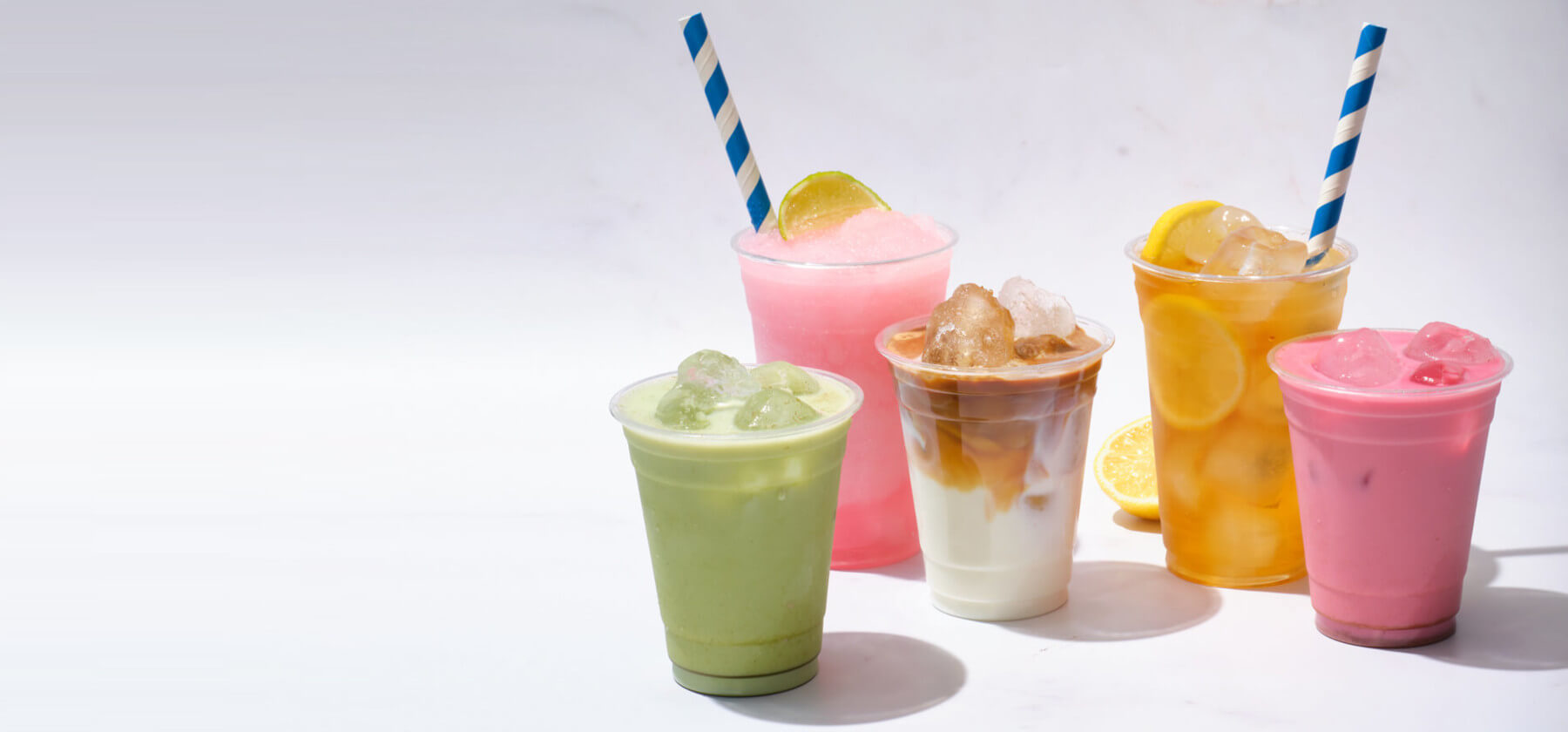 Five colourful iced drinks in plastic cups with straws, including pink slush, iced coffee, lemonade with lemon slices, green matcha, and pink milk tea, arranged on a white background.