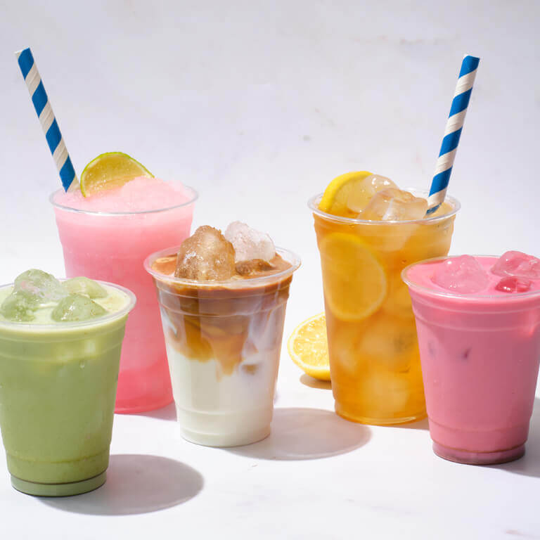 Five colourful iced drinks in plastic cups with straws, including pink slush, iced coffee, lemonade with lemon slices, green matcha, and pink milk tea, arranged on a white background.